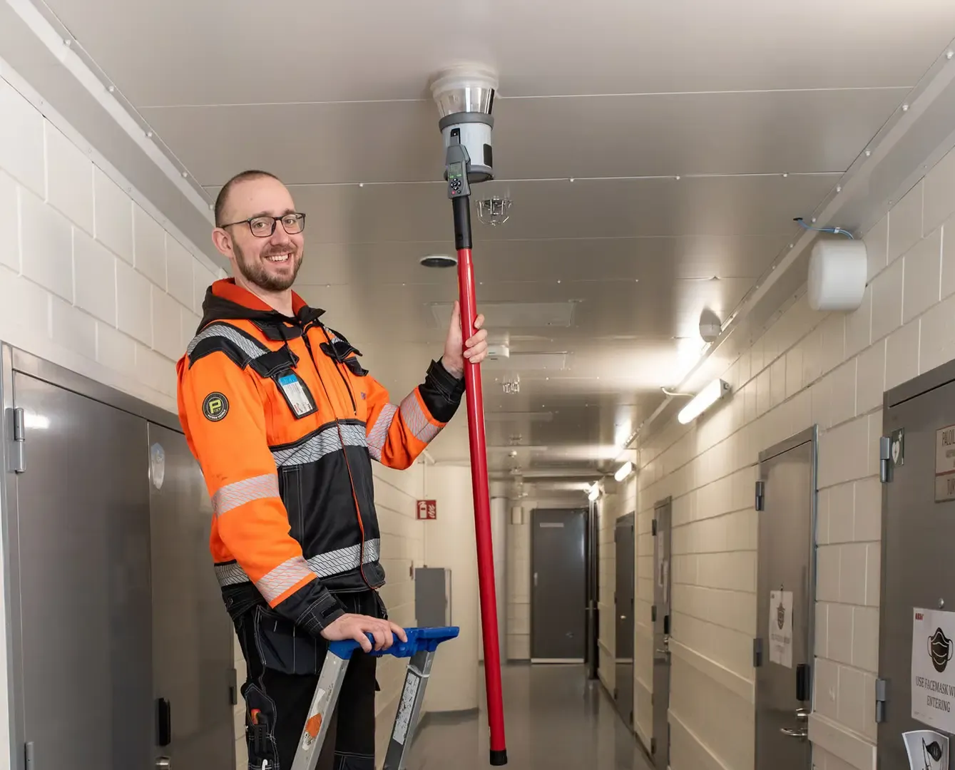 Oranssitakkinen asentaja testaa kattoilmaisinta pitkällä testivarrella käytävässä